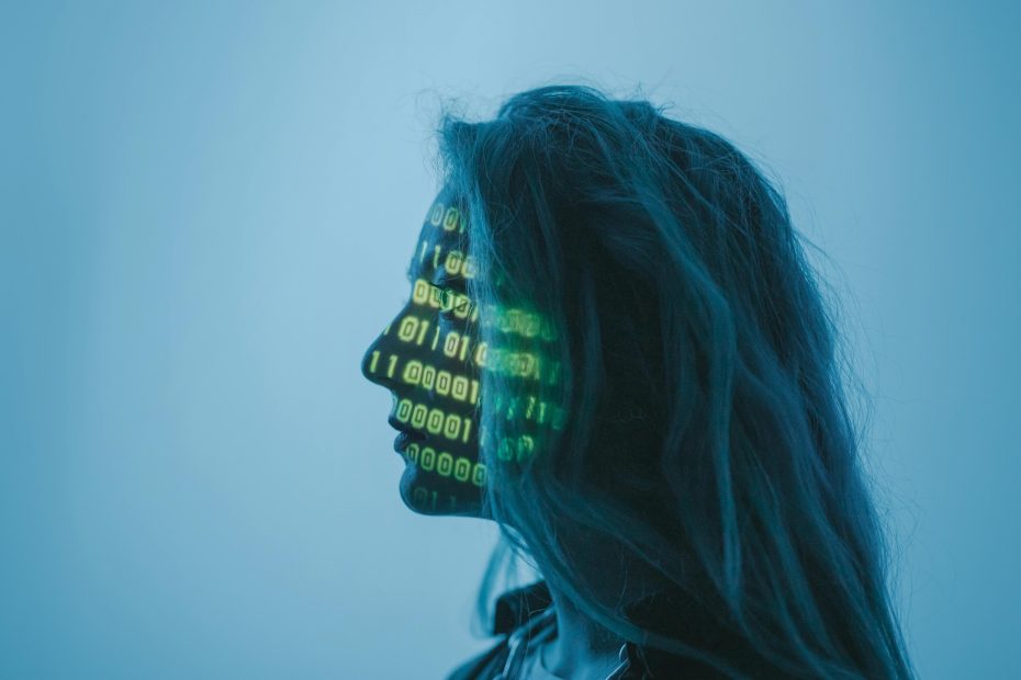 A profile silhouette of a person with long hair against a blue background. Bright green binary code numbers are projected onto their face, highlighting the eye and nose area, creating a digital or cybernetic aesthetic.