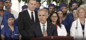 Prime Minister Mark Carney addresses a crowd at a press conference. Behind him are nurses/doctors in scrubs.